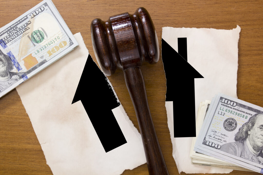An image showing a judge's gavel on a wooden surface next to a pile of $100 bills, with two large black arrows pointing upwards on torn paper. This suggests themes of rising financial stakes or costs, possibly in the context of legal proceedings or financial judgments.