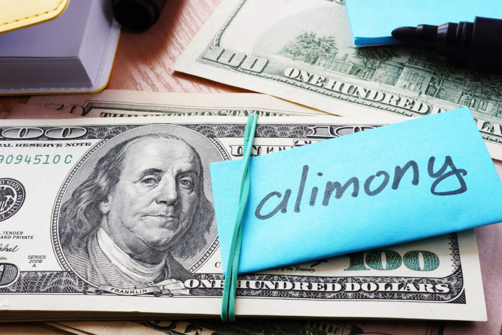 An image featuring a stack of $100 bills with a blue sticky note wrapped around them with the word 'alimony' written in black marker. In the background, there is a yellow legal pad and a black marker cap. The photo suggests the concept of spousal support payments in the context of family law.