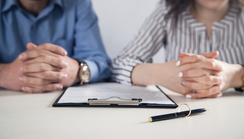 A focused image showing the clasped hands of a man and a woman seated at a table, symbolizing a collaborative or possibly meditative moment. Blurred in the foreground is a clipboard with documents, and a pen lying beside it, suggesting a formal meeting or legal procedure, potentially related to family law.